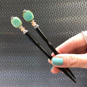 A hand holding a pair of the Dakota Tidal Hair Stick made from a turquoise Czech glass bead,