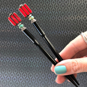 A hand holding two of the Nela Tidal Hair Sticks made from a red Czech glass bead.