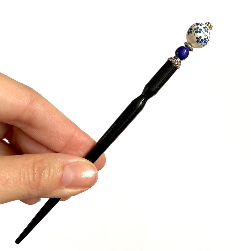 A hand holding two of the Nora Tidal Hair Sticks made from blue flowered white ceramic beads.
