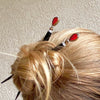 A woman wearing a hair bun using two of our Reina Hair Stick made from a red Czech glass bead with bronze edging.