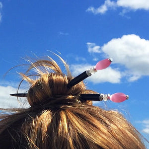 A woman wears a high bun with two of the Roxy Tidal Hair Sticks made of pink cat's eye glass.